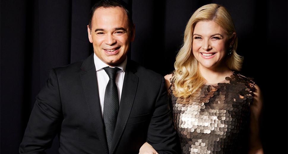 Male and woman performer smile at camera. Woman wears sparkly dress, man in black suit. 