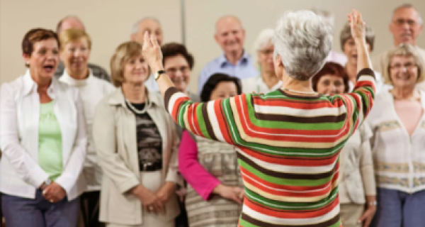 people lined up ready to sing in a choir. A lady is at the front with her arms in the air conducting the group.