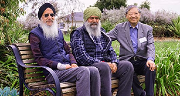 Three men sit on a park bench. Two have big white beards. 