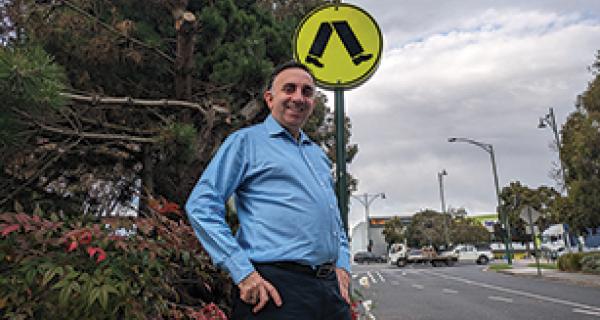 Man John in a blue shirt looks down at camera smiling, against Nunawading Civic Centre background of Whitehorse Road