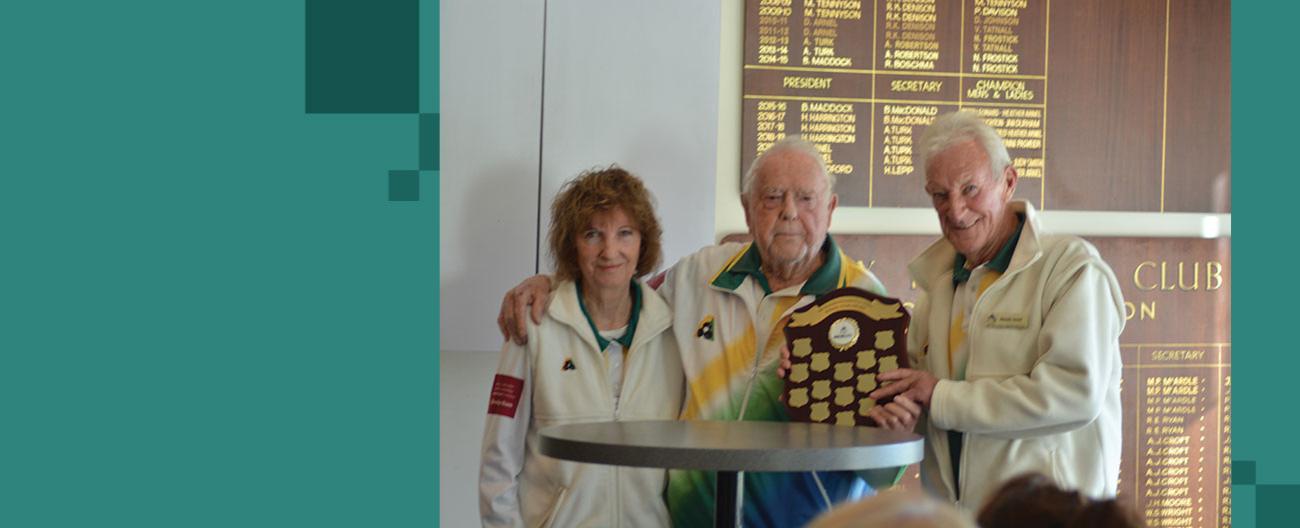 Man holds trophy up at bowl's club, supported by a man and woman on each side