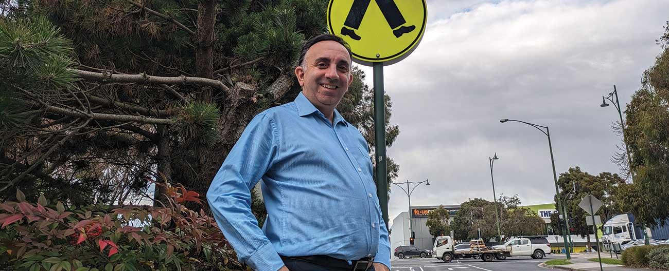 Man John in a blue shirt looks down at camera smiling, against Nunawading Civic Centre background of Whitehorse Road